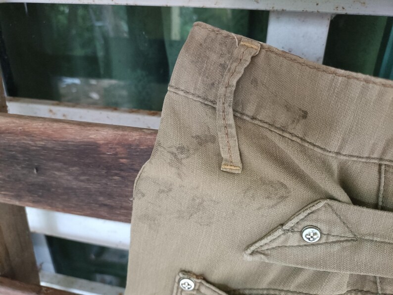 May include: Close-up of beige trousers showing wear and tear. The image focuses on the waistband, belt loops, and a pocket with a decorative button. The fabric shows signs of use, with stains and fading.