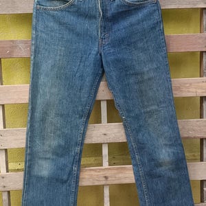 May include: Blue denim jeans with a classic five-pocket design. The jeans have a straight leg cut and a button and zipper closure. The denim shows some fading and wear, giving them a vintage look. The jeans are hanging on a black hanger.