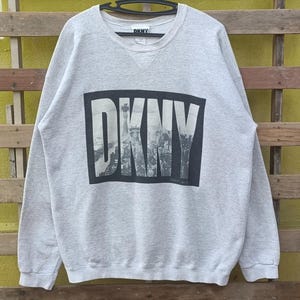 May include: A light gray DKNY sweatshirt with a crew neck. The front features a large black rectangle with the white DKNY logo and a grayscale image of a city skyline. The sweatshirt is hanging on a black hanger.