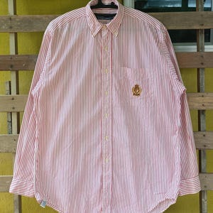 Puede incluir: Camisa abotonada de manga larga con rayas verticales rojas y blancas. La camisa tiene cuello abotonado, bolsillo en el pecho con emblema dorado y puños abotonados. La camisa está colgada de una percha negra.