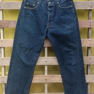May include: Dark blue denim jeans with a button fly and classic five-pocket design. The jeans are hanging on a black hanger against a yellow and wooden background. The jeans are a medium wash and appear to be vintage.