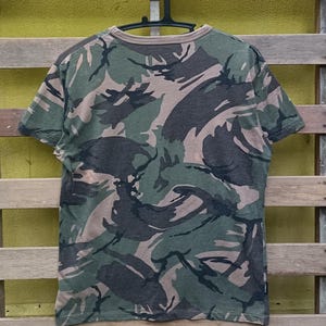 Puede incluir: Una camiseta de manga corta con estampado de camuflaje en tonos verdes, marrones y grises. La camiseta est&aacute; colgada de una percha negra sobre un fondo de madera. La parte trasera de la camiseta es visible, mostrando el estampado integral.