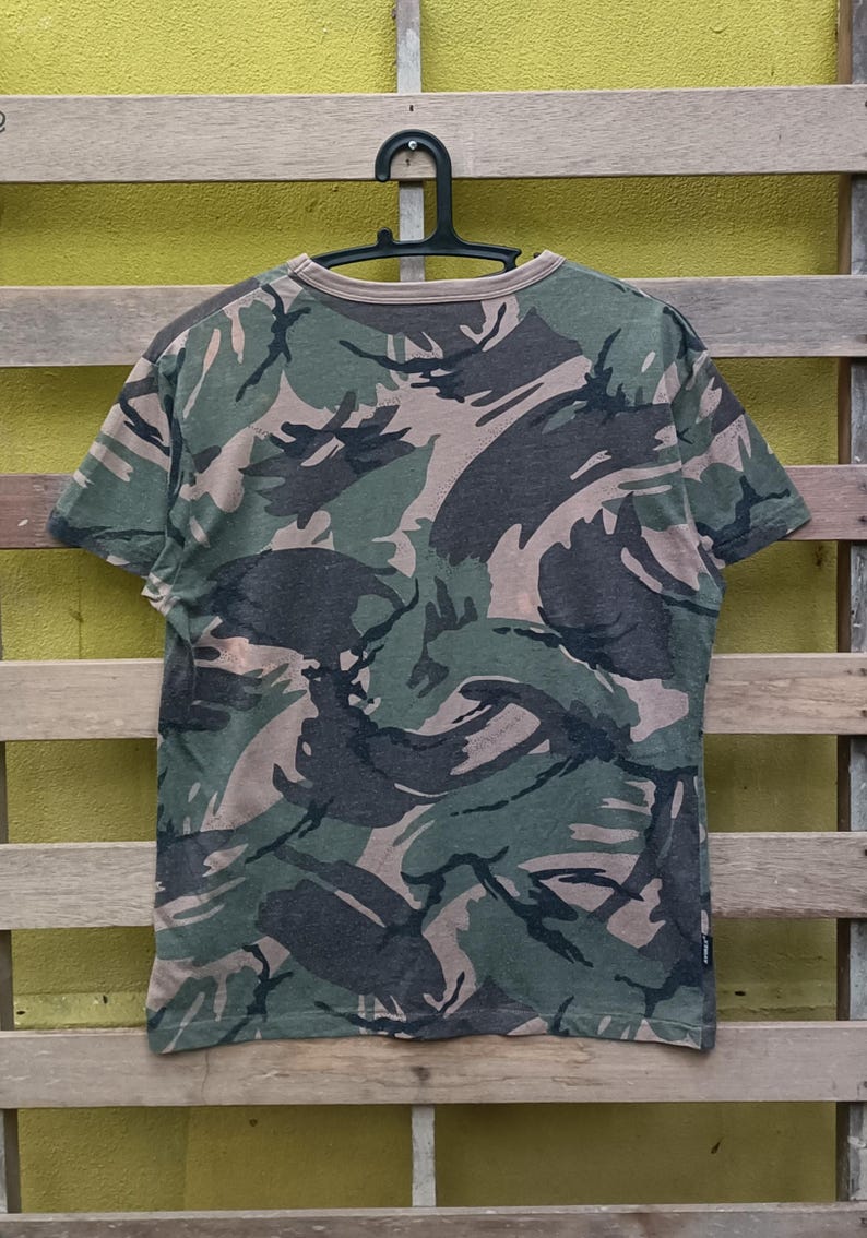 Puede incluir: Una camiseta de manga corta con estampado de camuflaje en tonos verdes, marrones y grises. La camiseta est&aacute; colgada de una percha negra sobre un fondo de madera. La parte trasera de la camiseta es visible, mostrando el estampado integral.