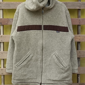 May include: A light gray fleece jacket with a hood and a full-length zipper. The jacket features a horizontal brown stripe across the chest and two front pockets. The jacket is displayed against a wooden background.
