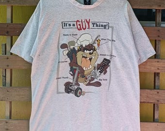 May include: A light gray t-shirt featuring a cartoon Taz character with the text "It's a GUY Thing!". The graphic includes golf clubs, a remote control, and other tools. The shirt has short sleeves and a crew neck.