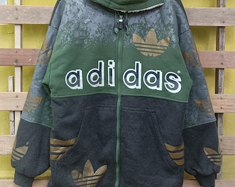 May include: A hooded zip-up jacket with the Adidas logo in gold and the brand name in white. The jacket features a color block design with green, gray, and black panels. The jacket has a fleece lining and two front pockets.