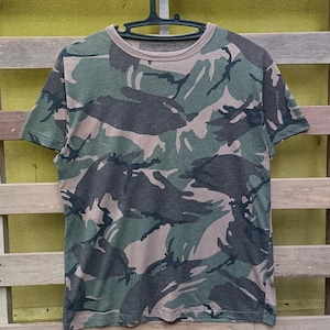 Puede incluir: Camiseta con estampado de camuflaje en tonos verdes, marrones y grises. La camiseta de manga corta est&aacute; colgada de una percha negra sobre un fondo de madera. El dise&ntilde;o de la camiseta presenta un estampado de camuflaje cl&aacute;sico de estilo militar.