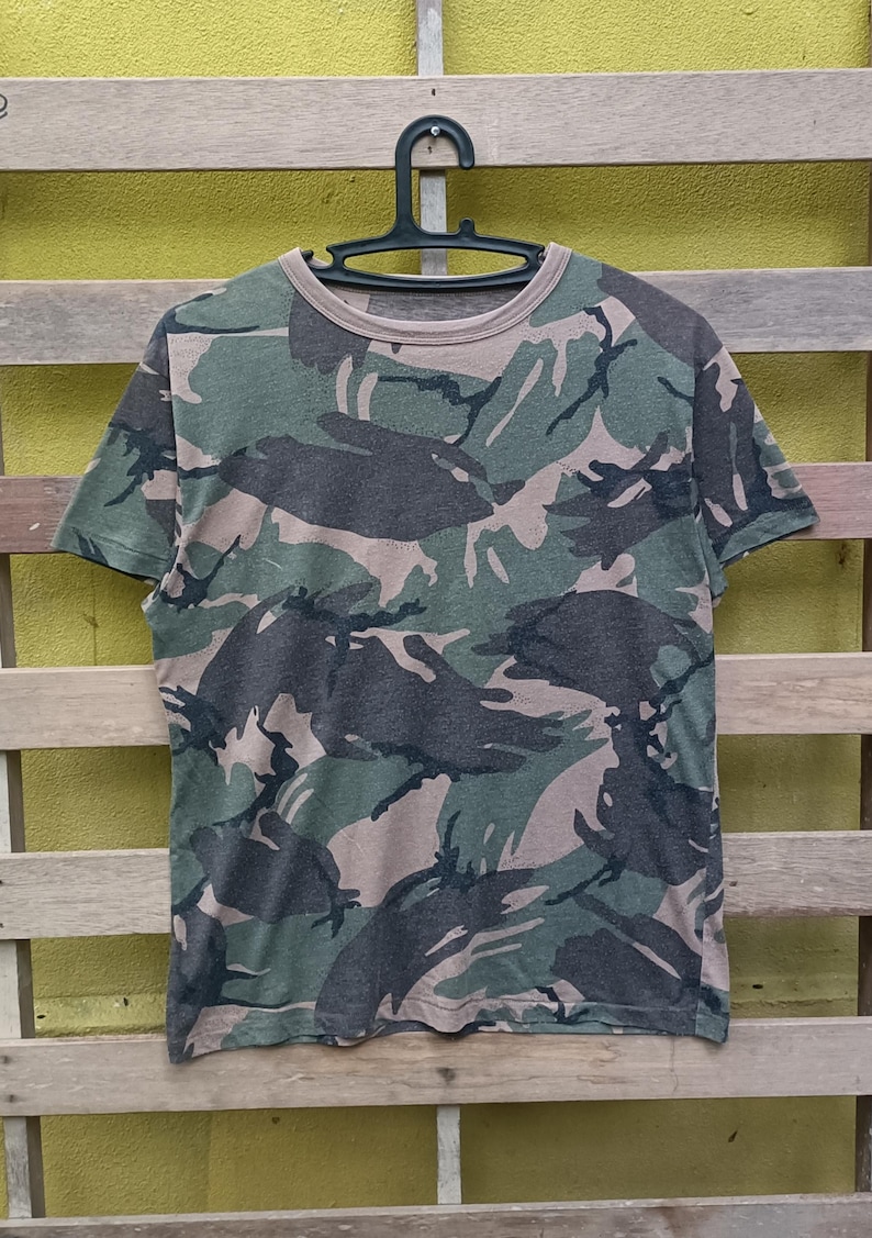 Puede incluir: Camiseta con estampado de camuflaje en tonos verdes, marrones y grises. La camiseta de manga corta est&aacute; colgada de una percha negra sobre un fondo de madera. El dise&ntilde;o de la camiseta presenta un estampado de camuflaje cl&aacute;sico de estilo militar.