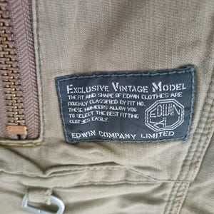 May include: A close-up shot of a khaki-coloured garment featuring a bronze zipper. A dark blue label displays "EXCLUSIVE VINTAGE MODEL" text regarding Edwin clothing fit and the Edwin logo.