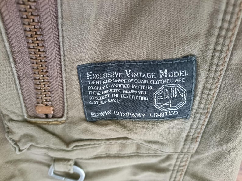 May include: A close-up shot of a khaki-coloured garment featuring a bronze zipper. A dark blue label displays "EXCLUSIVE VINTAGE MODEL" text regarding Edwin clothing fit and the Edwin logo.