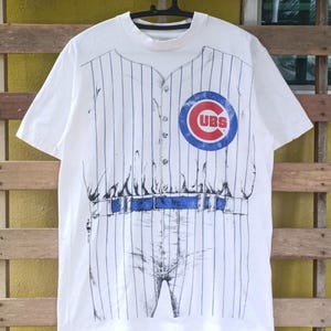 May include: White t-shirt featuring a baseball uniform design. The shirt has vertical blue stripes and a blue belt detail. The Chicago Cubs logo is on the chest. The shirt is short-sleeved and has a button-up front.