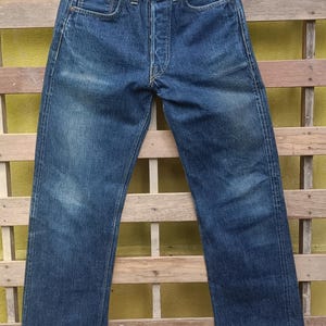 Può includere: Jeans di denim blu scuro appesi a una gruccia nera. I jeans hanno un classico design a cinque tasche, una chiusura con bottoni e un aspetto leggermente sbiadito. I jeans sono lunghi circa 81 cm.