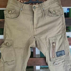 May include: Khaki cargo trousers with multiple pockets and a zip detail on the right leg. The trousers are hanging on a black hanger. The trousers have a button closure and a brown patterned interior waistband.