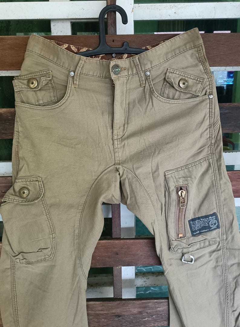 May include: Khaki cargo trousers with multiple pockets and a zip detail on the right leg. The trousers are hanging on a black hanger. The trousers have a button closure and a brown patterned interior waistband.