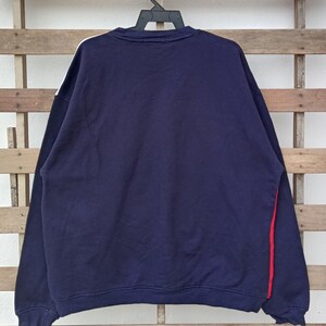 May include: Navy blue sweatshirt with white and red accents. The long-sleeved top features a white stripe on the left shoulder and a red stripe on the right side. The sweatshirt is hanging on a black hanger against a wooden background.