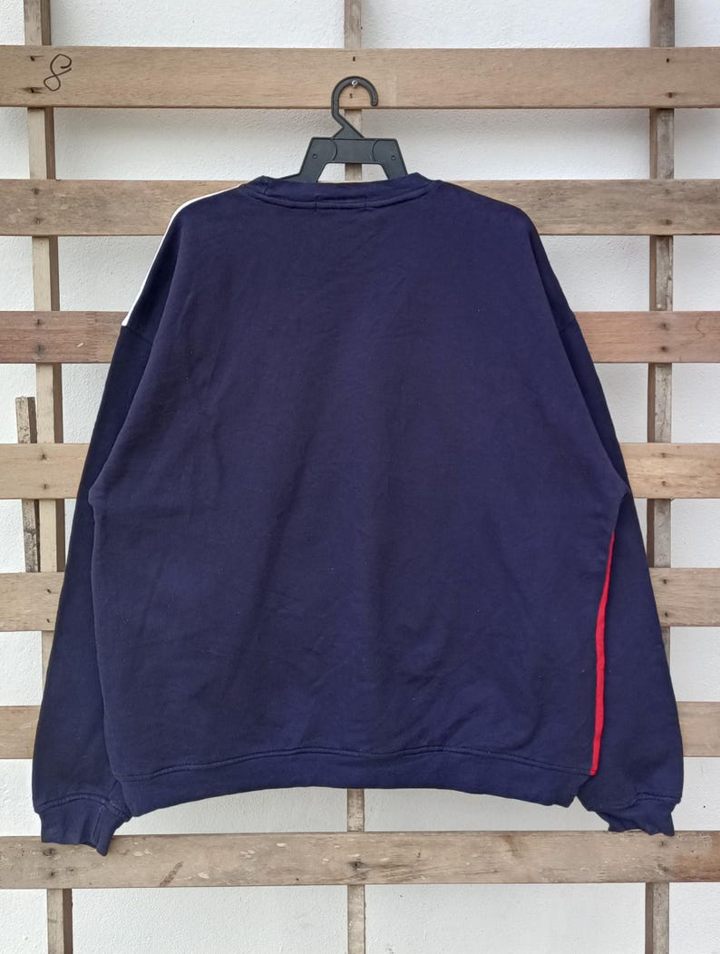 May include: Navy blue sweatshirt with white and red accents. The long-sleeved top features a white stripe on the left shoulder and a red stripe on the right side. The sweatshirt is hanging on a black hanger against a wooden background.