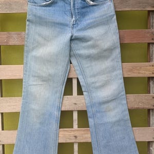 May include: Light blue denim jeans with a flared leg design, hanging on a black hanger. The jeans have a classic five-pocket design and a button and zipper closure. The denim has a faded appearance.