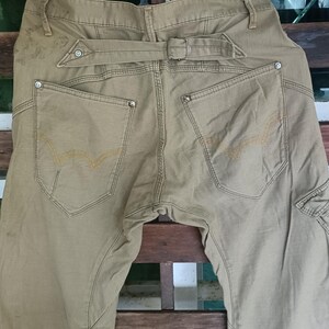 May include: Khaki-coloured trousers with a distinctive belt design and decorative stitching on the back pockets. The trousers have a cargo pocket on the side and are hanging on a black hanger.