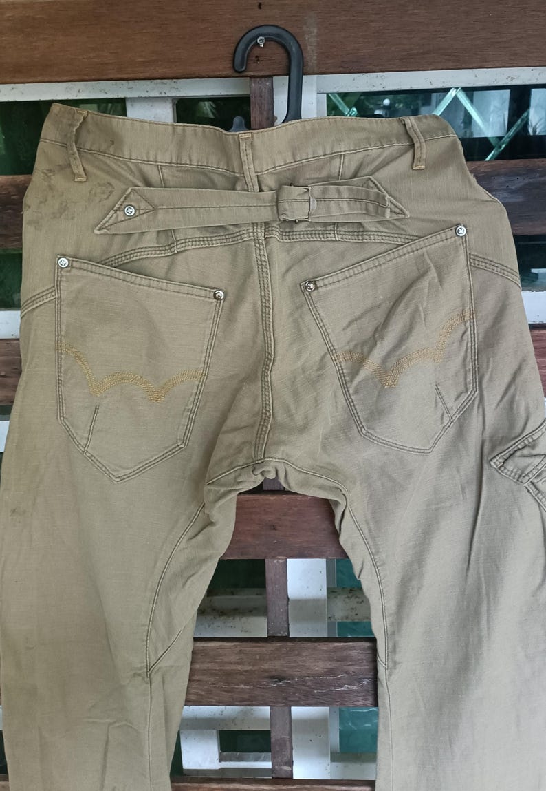 May include: Khaki-coloured trousers with a distinctive belt design and decorative stitching on the back pockets. The trousers have a cargo pocket on the side and are hanging on a black hanger.