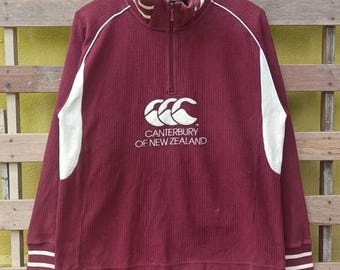 Puede incluir: Jersey de manga larga color burdeos con cuello con cremallera. Presenta detalles blancos en las mangas y el cuello, y el logotipo de Canterbury of New Zealand en el pecho. La tela acanalada tiene un aspecto texturizado. La prenda está colgada en una percha negra.