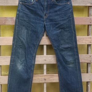 May include: Dark blue denim jeans hanging on a black hanger. The jeans have a classic five-pocket design with a button and zipper closure. The denim has some fading and wear, giving it a vintage look.