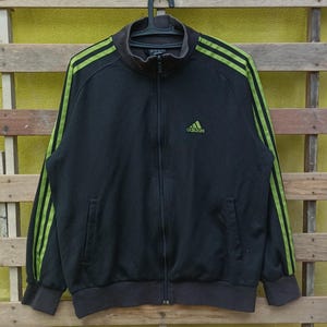 May include: Black zip-up track jacket with neon green stripes on the sleeves and the Adidas logo on the chest. The jacket has a high collar, two front pockets, and ribbed cuffs and hem. The jacket is hanging on a black hanger.