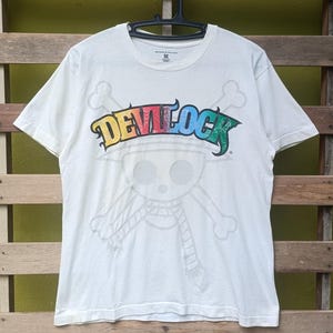 Puede incluir: Camiseta blanca con la palabra "DEVILOCK" en letras coloridas, un diseño de calavera y huesos cruzados, y un contorno de personaje de dibujos animados. La camiseta está colgada en una percha negra.