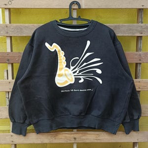 Puede incluir: Sudadera negra con un diseño de saxofón dorado y detalles blancos. El texto "devilock VS Garni desine wear" está impreso debajo. La sudadera está colgada en una percha negra sobre un fondo de madera amarilla y marrón.