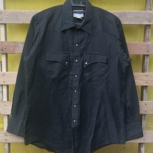 Op de afbeelding: Zwart shirt met lange mouwen en witte stikseldetails. Voorzien van twee borstzakken en een puntkraag. Het shirt is met knopen en heeft decoratieve stiksels langs de naden en manchetten. Het shirt hangt aan een zwarte hanger.