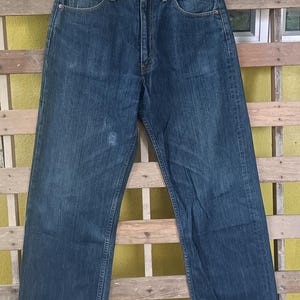 May include: Dark blue denim jeans with a classic five-pocket design. The jeans are hanging on a black hanger against a wooden background. The jeans have a straight leg cut.