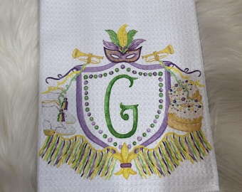 May include: White waffle-weave kitchen towel featuring a Mardi Gras crest design. The crest includes a purple and green shield with a large green letter "G", a mask, trumpets, and a cake. The towel has a decorative fringe.