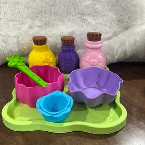 May include: A lime green tray holds three small bottles with brown caps, a bright pink bowl with a green flower-shaped spoon, a light purple bowl, and a small blue flower-shaped cup. The tray features embossed butterfly and flower designs.