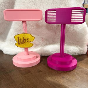 May include: Two plastic display stands with circular bases and vertical posts. The left stand is light pink with a rectangular top and a yellow, glittery, cloud-shaped ornament hanging from it that features the brown text "Lukes". The right stand is magenta.