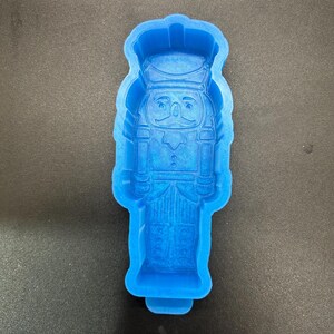May include: A bright blue silicone mold in the shape of a toy soldier nutcracker. The mold has detailed features, including a hat, uniform, and facial details. The mold is designed for creating decorative items or treats.