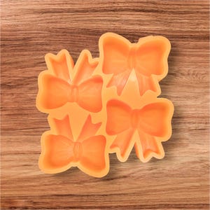 May include: Orange silicone mold with four bow shapes. The mold is designed for creating decorative bows for crafts or baking. The bows have detailed ribbon and knot designs. The mold is made of a flexible, food-safe material.