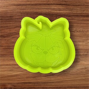 May include: A lime green silicone mold in the shape of a pumpkin with a witch hat and bow. The pumpkin has a face with large eyes and a mischievous grin, perfect for Halloween baking.