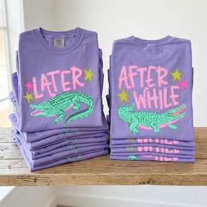 May include: Stacks of purple t-shirts with a graphic of a green alligator and the text "LATER" and "AFTER WHILE" in pink and yellow. The shirts are stacked on a wooden surface.