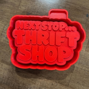 May include: A bright red cookie cutter with the words "NEXT STOP... THRIFT SHOP" in bold, raised letters. The cutter has a rectangular shape with rounded edges and a small tab at the top. The cutter is made of plastic.