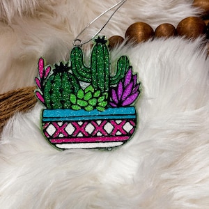 May include: A glittery cactus ornament featuring a variety of succulents in a blue, white, and pink patterned pot. The ornament has a silver wire for hanging. The cacti are green, pink, and purple.