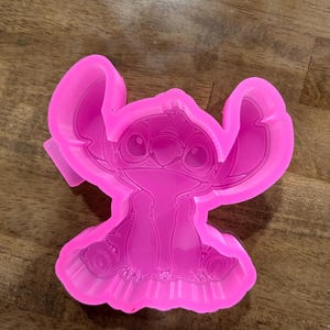 May include: A bright pink silicone mold in the shape of the character Stitch from the animated movie Lilo & Stitch. The mold features detailed facial features, large ears, and a textured surface, perfect for crafting projects.