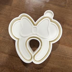 May include: A white plastic cookie cutter in the shape of a bow and mouse ears. The cutter has a rounded shape with a bow in the center and two circular shapes on either side, resembling mouse ears.