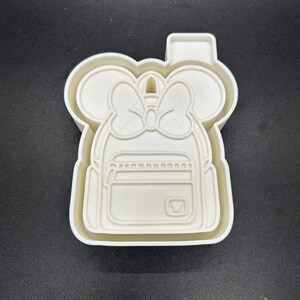 May include: A white plastic cookie cutter shaped like a backpack with mouse ears and a bow. The backpack features a front pocket and a small heart-shaped design. Ideal for making biscuits or other treats.