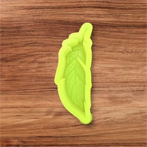 May include: A lime green silicone mold in the shape of a feather. The feather design has detailed patterns and textures. The mold is set against a brown wooden background.