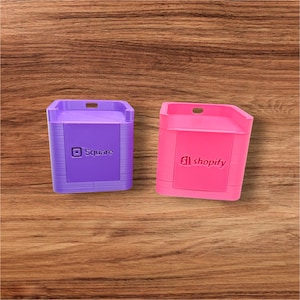 May include: Two small, rectangular containers, one purple with "Square" printed on it, and one pink with "Shopify" printed on it. Both have a small opening at the top and are set against a wooden background.