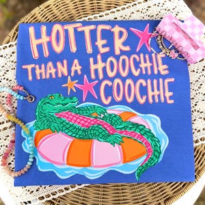 May include: A blue t-shirt with the text "HOTTER THAN A HOOCHIE COOCHIE" in pink and yellow. The shirt features a green alligator floating on a pink and orange inner tube. A pink star and a bracelet are visible.
