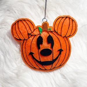 May include: An orange pumpkin-shaped ornament with black facial features and mouse ears. The ornament has a smiling face and a stem. The ornament is hanging from a silver metal loop.