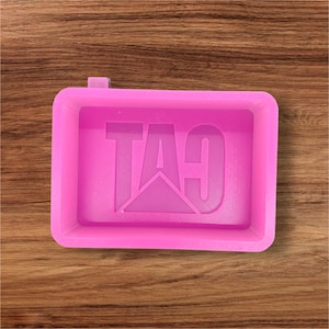 May include: A pink rectangular tray with the word "CAT" embossed in the center. The tray has a small tab on one side and is set against a wooden background. The CAT logo is a stylized design.