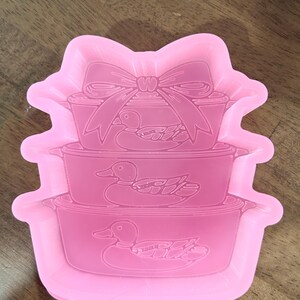 May include: Pink silicone mold featuring a stack of three containers with duck designs. The top container has a bow design. The mold is designed for crafting and creating decorative items. The background is a wooden surface.