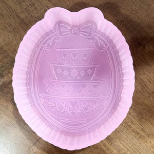 May include: A light pink silicone mold with a decorative design. The mold features an oval frame with a bow at the top and a stack of three bowls with floral and heart details. The outer edge has a ruffled border.