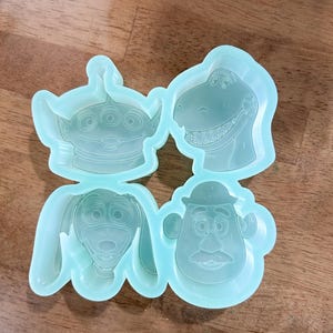 Peut inclure: Un moule en silicone bleu clair comportant quatre cavités de personnages distinctes. La cavité en haut à gauche représente un extraterrestre à trois yeux, celle en haut à droite un dinosaure souriant, en bas à gauche un chien aux longues oreilles.